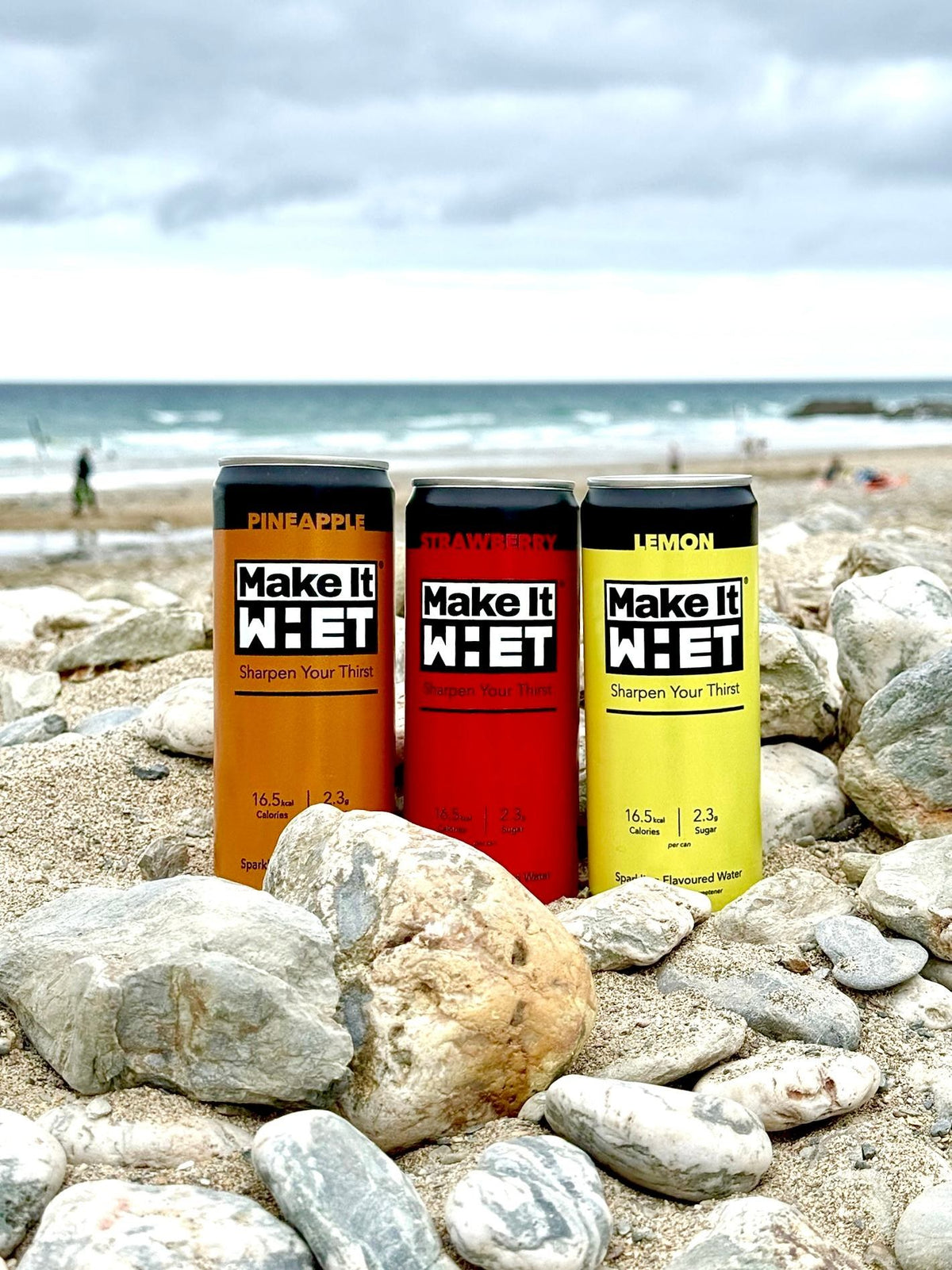 Three Make It WHET cans standing on pebbles beside the sea