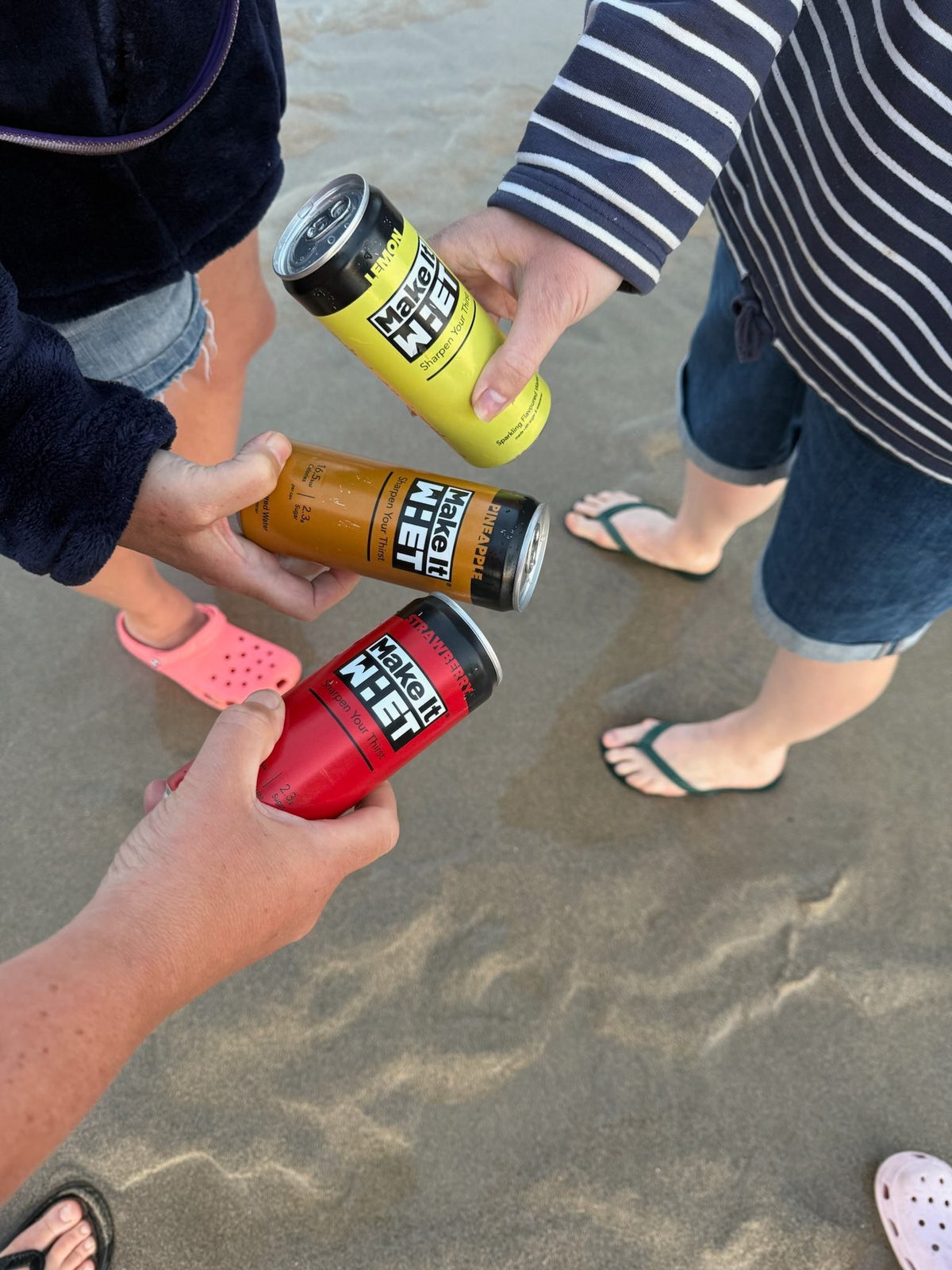 Three people holding Make It WHET cans together on a sandy beach