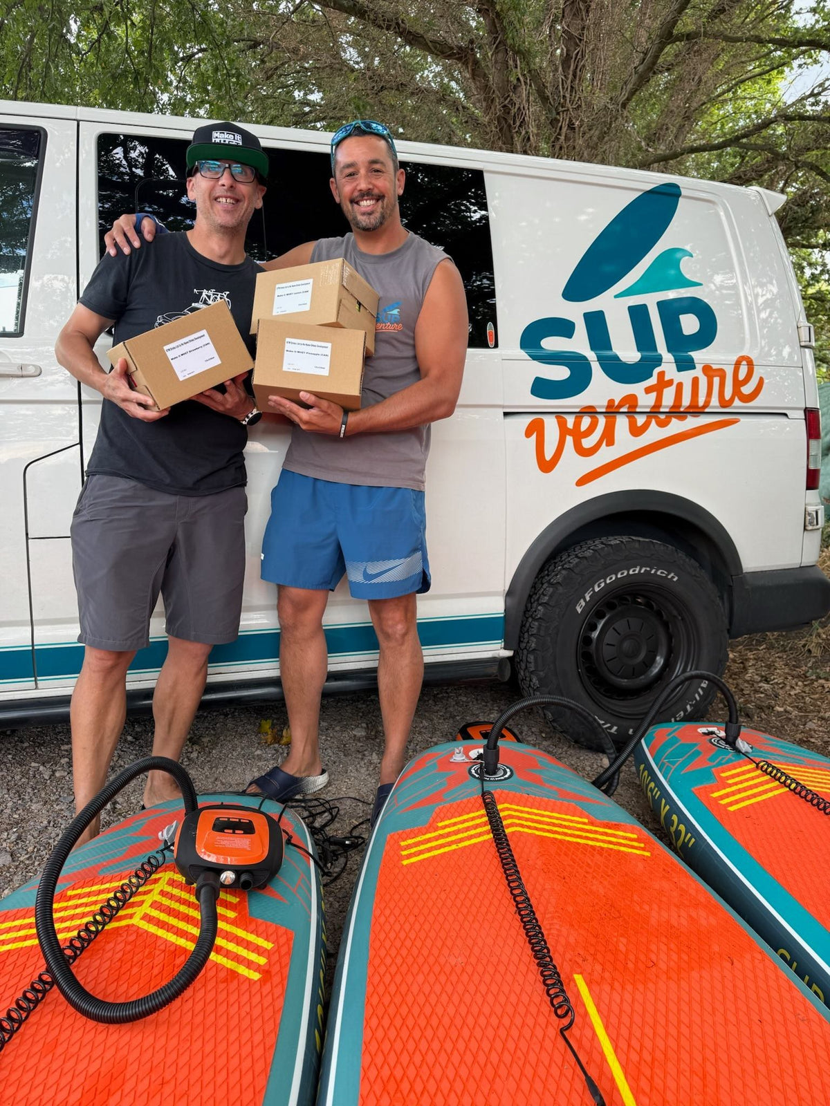 Two people holding boxes beside a SUP Venture van and paddleboards